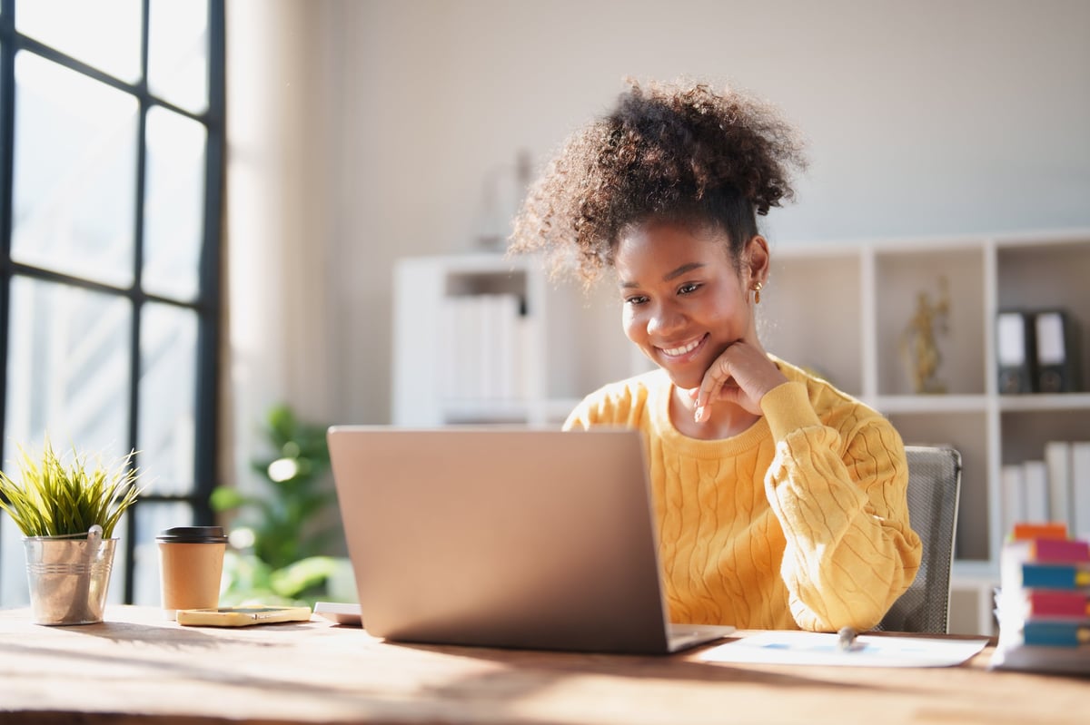 Young African American woman working from a bright home office, smiling at her laptop while managing her business